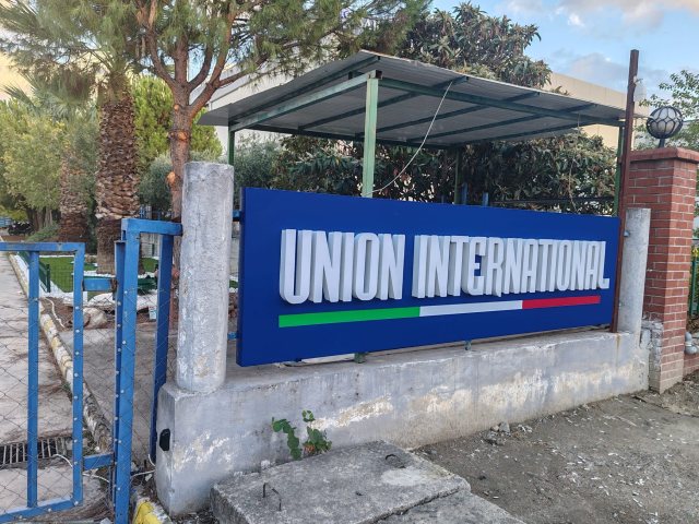 Union International fabrika girişinde mavi panolu, beyaz hacimli harfli tabela, gündüz